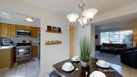 This two-story residence offers a thoughtfully arranged interior where key living spaces flow naturally into one another. The dining area serves as a central hub, anchored by a dark wood dining table set beneath a brushed nickel three-arm chandelier that provides warm, focused lighting. Decorative floating shelves along the dining wall add a personalized touch, displaying small framed artwork and ornamental accents.

Adjacent to the dining space, the kitchen is outfitted with oak cabinetry, tile flooring, and stainless steel appliances including a refrigerator and built-in microwave, offering a practical and efficient cooking environment. A tiled entryway with a utility mat marks the transition into this culinary space.

Beyond the dining area, the carpeted living room receives natural light through a window fitted with horizontal blinds, creating a relaxed and comfortable atmosphere. The open sightlines between the dining room, kitchen, and living area contribute to a cohesive, connected feel throughout the main level. The two-story layout suggests additional living or sleeping quarters above, expanding the overall functionality of the home.