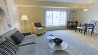 This single-floor space offers a warm and inviting atmosphere defined by its thoughtful design and neutral color palette. The living area serves as the heart of the space, featuring soft yellow walls that lend a cheerful, airy quality to the interior. A large picture window takes center stage, flooding the room with abundant natural light and providing a pleasant view of the outdoors. The flooring transitions seamlessly between light hardwood and a plush area rug, creating distinct zones for relaxation and conversation. Comfortable seating, including a gray sofa and a coordinating upholstered armchair, are arranged to encourage a relaxed and social atmosphere. Nesting round coffee tables anchor the seating area, adding a modern touch to the overall design. Decorative accents, including a patterned wall piece and a sleek console table along the wall, contribute to the curated yet approachable aesthetic. A tall floor lamp provides warm ambient lighting, complementing the natural light from the large window. The overall ambiance is clean, calm, and functional, making this a versatile space well-suited for both everyday living and entertaining.
