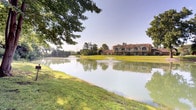 This single-story clubhouse is situated on a beautifully landscaped property bordering a tranquil pond adorned with decorative fountains. Mature trees frame the exterior, which features a distinctive pitched roofline with gabled accents and warm brick façade elements that complement the natural surroundings.

Inside, the main lounge area is defined by soaring vaulted ceilings with exposed beam detailing and ceiling fans, creating an airy, open atmosphere. A floor-to-ceiling brick fireplace serves as a prominent focal point, adding warmth and architectural character to the space. Plush sofas and upholstered armchairs are arranged throughout, alongside glass-topped coffee tables, providing comfortable seating for gatherings. Recessed lighting and large windows with vertical blinds allow generous natural light to filter throughout.

Adjacent to the lounge, a bar area features a tiled countertop with bar stool seating, a granite high-top table, and access to a kitchenette equipped with a refrigerator. Sliding glass doors open toward an outdoor pool area, enhancing the connection between interior and exterior spaces. The reception area near the entrance includes additional seating and a welcoming, well-appointed layout suited for community use.