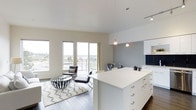 This contemporary single-floor space showcases a thoughtfully designed open-concept layout that seamlessly connects the kitchen, dining, and living areas into one cohesive environment. The kitchen is a standout feature, anchored by a large white island with ample storage, crisp white cabinetry, and a striking navy tile backsplash. Pendant lighting hangs above the island, adding both function and a modern design touch. The living area is bathed in natural light, courtesy of floor-to-ceiling windows that frame sweeping panoramic views of the surrounding cityscape and horizon. Light wood-toned flooring runs throughout, complementing the neutral white and soft grey color palette that defines the interior. The living space is open and airy, with high ceilings that enhance the sense of volume and flow. A designated dining area with modern seating bridges the kitchen and living zones effortlessly. Track lighting and recessed fixtures provide additional illumination throughout. The overall ambiance is clean, bright, and metropolitan — an ideal blend of modern functionality and contemporary design that takes full advantage of its elevated position and expansive natural light.