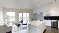 This contemporary single-floor space showcases a thoughtfully designed open-concept layout that seamlessly connects the kitchen, dining, and living areas into one cohesive environment. The kitchen is a standout feature, anchored by a large white island with ample storage, crisp white cabinetry, and a striking navy tile backsplash. Pendant lighting hangs above the island, adding both function and a modern design touch. The living area is bathed in natural light, courtesy of floor-to-ceiling windows that frame sweeping panoramic views of the surrounding cityscape and horizon. Light wood-toned flooring runs throughout, complementing the neutral white and soft grey color palette that defines the interior. The living space is open and airy, with high ceilings that enhance the sense of volume and flow. A designated dining area with modern seating bridges the kitchen and living zones effortlessly. Track lighting and recessed fixtures provide additional illumination throughout. The overall ambiance is clean, bright, and metropolitan — an ideal blend of modern functionality and contemporary design that takes full advantage of its elevated position and expansive natural light.