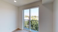 This single-floor space showcases a clean, modern design defined by minimalist finishes and an abundance of natural light. The featured room is anchored by large sliding glass doors framed in white, which open directly onto a private balcony enclosed by sleek black metal railings. The elevated position of the balcony affords expansive views of lush tree canopies, neighboring rooftops, and a distant city skyline, creating a strong visual connection between the interior and the outdoors. The room's interior is finished with crisp white walls and smooth light wood flooring, contributing to an airy, contemporary atmosphere. Recessed lighting in the ceiling supplements the generous daylight streaming through the glass doors. The overall aesthetic is understated and functional, with clean lines and neutral tones throughout. The balcony access enhances the sense of openness, making the space feel larger than its footprint suggests. This room is well-suited as a bedroom or living area, benefiting from both privacy and a compelling outdoor perspective that brings natural elements into the everyday environment.
