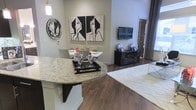 This single-floor residence showcases a thoughtfully designed open-concept layout that seamlessly connects the kitchen, dining, and living areas. The kitchen features a striking granite-topped island with elegant curved edges, providing both a functional workspace and a natural gathering point. Rich, dark hardwood flooring runs throughout the main living areas, adding warmth and sophistication to the space. The dining area is anchored by a clean white table surrounded by modern chairs, illuminated by a sleek pendant light overhead. Bold black-and-white artwork adorns the neutral gray walls, lending the space a contemporary, gallery-like ambiance. The living area is defined by a plush white area rug with a subtle geometric pattern, complementing the overall monochromatic design palette. A dark media console serves as the focal point of the living room, topped with decorative accents that echo the space's modern aesthetic. A wall-mounted flat-screen television completes the entertainment setup. Natural light filters in through draped windows, further brightening the open interior. The combination of granite surfaces, hardwood floors, and curated art creates a cohesive, upscale atmosphere throughout this well-appointed single-level residence.
