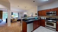 This single-floor residence offers a thoughtfully designed open-concept layout that seamlessly connects the kitchen and living area. The kitchen is outfitted with rich cherry wood cabinetry, dark granite countertops, stainless steel appliances including a gas range with over-the-range microwave, a dishwasher, and a double-basin sink. A peninsula with pendant chandelier lighting serves as a natural divider between the kitchen and the living area, which features soft carpeting, neutral walls, and a large window that allows natural light to fill the space. The living room provides a comfortable, open atmosphere with generous wall space for artwork display. The bedroom is carpeted and well-proportioned, featuring neutral tones, ample room for furnishings, and access to a walk-in closet area. The full bathroom includes a cherry wood vanity with a single sink, a large mirror with overhead lighting, tile flooring, and a shower-tub combination. A dedicated in-unit laundry room houses a full-size washer and dryer with overhead shelving, adding everyday convenience. Throughout the residence, consistent use of tile and carpet flooring, warm cabinetry tones, and neutral wall colors create a cohesive and functional living environment.