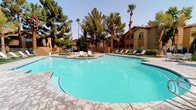 This inviting community showcases a distinctive Southwest-inspired architectural style, characterized by warm stucco exteriors, red tile roofing, and arched windows that lend the property a Mediterranean charm. The centerpiece of the outdoor space is a generously sized, freeform swimming pool with vivid turquoise water, surrounded by a stamped concrete deck ideal for lounging. Ample poolside seating, including lounge chairs and shaded umbrella tables, provides comfortable spaces for relaxation and socializing. A separate spa offers a more intimate retreat, complete with stainless steel entry rails and bordered by manicured landscaping. Hammocks and green turf areas add a casual, recreational touch to the outdoor environment. Mature palm trees, pines, and lush desert-adapted plantings create a shaded, resort-like ambiance throughout the grounds. The surrounding multi-story buildings frame the pool courtyard, contributing to a sense of privacy and community. Inside, the space includes an accessible shower room finished with large-format, light-toned wall tiles and small mosaic floor tiles in a neutral tone. Stainless steel grab bars, a handheld showerhead, and dual fixed rain-style showerheads ensure both functionality and accessibility. Overall, the space combines practical amenities with an appealing outdoor lifestyle aesthetic.