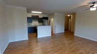 This single-floor space is designed with an open-concept layout that integrates the kitchen and living areas into one cohesive, functional environment. The kitchen is outfitted with dark espresso cabinetry that provides a striking contrast against the light-toned walls and flooring. A peninsula with a white base serves as a natural divider between the kitchen and the living area, offering additional counter space and a casual gathering spot. Stone-look countertops and a stainless steel refrigerator contribute a modern, polished aesthetic to the cooking space, which also includes a range and an overhead microwave or cabinet unit. A flush-mount fluorescent light fixture illuminates the kitchen area effectively. Warm hardwood-style flooring extends throughout the open living space, adding continuity and a welcoming feel underfoot. Crown molding along the upper walls adds a subtle architectural detail to the interior. Hallways visible toward the rear of the unit lead to additional rooms, suggesting a well-organized floor plan. The overall ambiance is clean, functional, and move-in ready, with neutral tones providing a versatile backdrop.