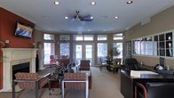 This single-floor space serves as an inviting reception and communal lounge, thoughtfully designed to accommodate both relaxation and social interaction. The curved architectural layout lends a sense of openness, while a bank of large windows and glass-paneled French doors wraps around the room, flooding the interior with generous natural light and offering views of the surrounding greenery outside.

The ceiling is finished with recessed lighting and crown molding, complemented by a centrally mounted ceiling fan that adds both function and a decorative touch. The flooring transitions between carpet and hard surface, subtly defining distinct zones within the space.

Seating throughout the lounge is varied and plentiful, including cushioned armchairs, sofas, and dining-style chairs arranged around low coffee tables and accent tables. Decorative potted plants and floral arrangements add warmth and a natural element to the interior. Along one side of the room, a reception or service desk is positioned near display cabinetry, suggesting a staffed welcome area.

A separate dining or activity zone is visible toward the rear, furnished with tables and chairs for group use. The overall ambiance is warm, well-lit, and professionally appointed, making this space well-suited for a residential clubhouse, leasing office, or community gathering area.