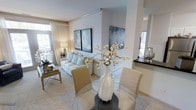 This single-floor, 2-bedroom, 2-bathroom residence features an open-concept design that connects the living, dining, and kitchen areas into one cohesive, light-filled space. The living area is highlighted by soft neutral tones, plush carpeting, and crown molding details that lend a refined finish to the interior. Large windows dressed with sheer draping allow generous natural light to filter throughout, creating a bright and airy ambiance.

The dining area sits adjacent to the living space, accommodating a glass-top table with upholstered seating — an ideal arrangement for both casual meals and entertaining. A partial wall with a dark countertop surface separates and subtly defines the kitchen zone, which is visible in the background and appears to feature sleek finishes.

Curated décor, including framed artwork and a sculptural floral arrangement, contributes to the space's polished, contemporary aesthetic. The overall color palette of whites, grays, and soft blues creates a calm and sophisticated atmosphere throughout. The thoughtful layout maximizes the sense of openness while maintaining defined functional areas, making this residence both practical and visually appealing.