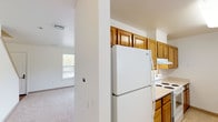 This two-floor residential space offers a practical and well-organized layout designed for comfortable everyday living. The kitchen is efficiently arranged in a galley style, featuring warm-toned oak cabinetry that extends to the upper walls for ample storage, light-colored countertops, and white appliances including a refrigerator and electric range. The tile flooring adds durability to the space. Adjacent to the kitchen, a bright living and dining area benefits from multiple large windows fitted with blinds, allowing generous natural light to fill the room. A pendant light fixture adds a finishing touch overhead, and soft carpeting runs throughout the common and bedroom areas. The bedrooms are generously sized with neutral walls, carpeted floors, built-in closets, and windows that frame outdoor greenery views. One bedroom includes a walk-in style closet. The space includes two bathrooms: one full bath with a single vanity sink, mirror with overhead lighting, and a shower/tub combination; and a second bathroom thoughtfully equipped with accessibility features, including grab bars along the walls and an open-clearance vanity. The overall design is clean and neutral, creating a bright, move-in-ready environment with a practical, no-frills aesthetic suitable for a variety of occupants.