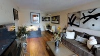 This single-floor space showcases an open-concept layout that connects the living and dining areas in a cohesive, functional arrangement. Rich hardwood flooring runs throughout, adding warmth and continuity to the space. The living area is anchored by a plush grey sofa accessorized with textured throw pillows, paired with a sturdy metal-framed coffee table that adds an industrial touch. A bold abstract wall piece serves as a striking focal point, complementing the curated artwork displayed across the walls.

The dining area, positioned just beyond the living space, features a dark wood table surrounded by white upholstered chairs, topped by a ceiling fan that provides both airflow and a subtle design element. A low dark credenza with decorative accents, small sculptures, and greenery adds depth and visual interest to the shared space.

Natural light filters through the windows, brightening the neutral-toned walls and highlighting the warm tones of the hardwood floors. Lush indoor plants are thoughtfully placed throughout, enhancing the inviting ambiance. The textured ceiling adds architectural character, while the overall design reflects a balanced blend of contemporary style and comfortable livability.