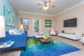 This single-floor residence is thoughtfully designed with a cohesive color palette and distinct character in each room. The living area serves as the heart of the space, featuring a bold teal accent wall, crown molding, plush carpeting, and a ceiling fan overhead. Natural light filters in through windows with blinds and a glass-paneled door leading to the exterior, creating a bright and welcoming atmosphere. The room accommodates generous seating and a central coffee table, making it well-suited for both relaxing and entertaining.

Adjacent to the living area, the kitchen is equipped with light wood cabinetry, dark granite-style countertops, tile flooring, and black appliances. A peninsula with bar seating connects the kitchen to a dining area, enhancing the open flow between spaces. Crown-molded columns add an architectural accent at the transition point.

The bedrooms feature carpeted floors, ceiling fans, and crown molding. One bedroom is distinguished by a vibrant yellow accent wall, teal curtains framing a window with wooded views, and ample space for a full bedroom set. Another bedroom includes a walk-in closet and a large framed mirror above a wood dresser, adding warmth and functionality. Throughout, the residence maintains a consistent blend of colorful accents and classic architectural detailing.