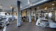 Located at 2170 Benton Blvd in Savannah, Georgia, this fitness center offers a well-equipped, single-floor workout facility designed to accommodate a wide range of exercise needs. The space is defined by its industrial-inspired aesthetic, featuring exposed spiral ductwork running along the ceiling, recessed and track lighting, and bold dark support columns that frame the open floor plan. Durable rubber flooring throughout provides both safety and comfort during workouts. The facility is outfitted with a comprehensive selection of strength training machines and cardio equipment, including stair climbers, cable machines, and seated resistance stations. Full-length mirrors line the walls, enhancing the sense of space and allowing users to monitor their form. The open layout ensures ample room to move between equipment, while the neutral gray color palette and modern finishes give the space a clean, professional atmosphere. Ventilation is well-integrated into the design, contributing to a comfortable environment during exercise. Overall, this fitness center combines functional design with a modern industrial character, making it a practical and well-appointed facility for residents or members seeking a comprehensive on-site workout space.