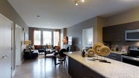 This single-floor residence is designed around an open-concept layout that seamlessly connects the kitchen, dining, and living areas into one cohesive, flowing space. The kitchen anchors the layout with a generously sized granite-topped island, offering both prep space and bar-style seating with sleek modern stools. A stainless steel faucet and contemporary decorative accents complete the polished kitchen aesthetic.

The living area extends naturally from the kitchen, furnished with dark upholstered seating accented by rust and orange throw pillows that add warmth against the cool grey walls. Large windows dressed with burnt orange curtains allow substantial natural light to filter through, creating a bright and inviting atmosphere. Track lighting overhead supplements the natural light and highlights the clean, modern ceiling.

A defined dining area sits between the kitchen and living room, featuring a dark table surrounded by matching chairs, maintaining visual consistency throughout the open floor plan. An adjacent doorway leads to a bedroom, visible in the background with warm lamp lighting, offering a private retreat from the main living space. The overall ambiance is contemporary and cohesive, with a refined neutral palette punctuated by rich accent tones.