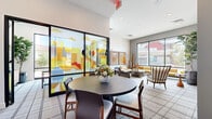 Located at 9100 S 59th Ave in Phoenix, AZ, this single-floor community lounge space offers a thoughtfully designed interior that blends contemporary aesthetics with comfortable functionality. The space is anchored by soaring ceilings that amplify the sense of openness, while large floor-to-ceiling windows and glass panel walls flood the interior with abundant natural light and offer views of the surrounding outdoor landscape.

A striking, colorful cityscape mural on the glass partition serves as a bold focal point, infusing the space with artistic energy. The lounge is divided into distinct yet connected zones: a dining area centered around a round dark-wood table with upholstered chairs, and a relaxed seating area featuring a sleek mustard-yellow sofa, a round concrete coffee table, and accent chairs. Curated artwork — including abstract paintings and framed prints — adorns the walls, complementing the warm color palette of yellows, oranges, and earthy tones throughout.

A herringbone-patterned wooden credenza and decorative sculptural pieces add refined, textural contrast to the space. The plaid-patterned carpet ties the zones together, while lush indoor plants, including a large fiddle-leaf fig, introduce a natural, organic element. Recessed lighting and natural light work in harmony to create a bright, inviting ambiance throughout.