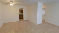 This single-floor space is designed with a straightforward, functional layout that maximizes usability. The main living area is finished with light beige carpeting and cream-colored walls, creating a neutral and versatile backdrop suitable for a variety of furnishing styles. A structural column adds a subtle architectural element to the open space, partially dividing the room while maintaining an open feel.

An open doorway connects the living area directly to the kitchen, which features warm-toned wooden cabinetry along the upper and lower sections of the walls. The kitchen is finished with light-colored countertops and tile flooring, offering a practical contrast to the carpeted living space. The cabinetry provides ample storage, and the layout appears efficient for everyday use.

A ceiling fan is visible in the living area, contributing to climate comfort. Additional doorways suggest further rooms or hallways beyond the main living space. Overall, the unit presents a clean, move-in-ready environment with a simple and cohesive design aesthetic throughout.
