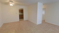 This single-floor space is designed with a straightforward, functional layout that maximizes usability. The main living area is finished with light beige carpeting and cream-colored walls, creating a neutral and versatile backdrop suitable for a variety of furnishing styles. A structural column adds a subtle architectural element to the open space, partially dividing the room while maintaining an open feel.

An open doorway connects the living area directly to the kitchen, which features warm-toned wooden cabinetry along the upper and lower sections of the walls. The kitchen is finished with light-colored countertops and tile flooring, offering a practical contrast to the carpeted living space. The cabinetry provides ample storage, and the layout appears efficient for everyday use.

A ceiling fan is visible in the living area, contributing to climate comfort. Additional doorways suggest further rooms or hallways beyond the main living space. Overall, the unit presents a clean, move-in-ready environment with a simple and cohesive design aesthetic throughout.
