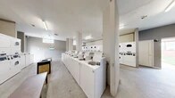 Located at 7003 Dunmanway in Dundalk, Maryland, this shared laundry facility offers a practical and efficiently organized space for residents. The single-floor layout makes excellent use of its footprint, accommodating a generous number of commercial-grade, top-load washing machines arranged in a long row down the center of the room, complemented by a bank of stacked dryers along the perimeter and in a dedicated alcove. The interior features concrete flooring, structural columns, and neutral gray and white walls that give the space a clean, utilitarian character. Overhead fluorescent lighting ensures the entire area is well-illuminated throughout, while glass block windows along the upper walls allow additional natural light to filter in. A centrally placed bench provides a convenient spot for folding or waiting. The facility is accessible via a door that opens to the exterior, allowing for easy entry and exit. The layout is open and unobstructed, enabling multiple users to move freely between machines without congestion. Overall, this laundry facility is designed with functionality and efficiency in mind, providing residents with a well-equipped communal space to meet their laundry needs.