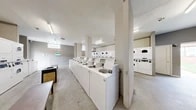 Located at 7003 Dunmanway in Dundalk, Maryland, this shared laundry facility offers a practical and efficiently organized space for residents. The single-floor layout makes excellent use of its footprint, accommodating a generous number of commercial-grade, top-load washing machines arranged in a long row down the center of the room, complemented by a bank of stacked dryers along the perimeter and in a dedicated alcove. The interior features concrete flooring, structural columns, and neutral gray and white walls that give the space a clean, utilitarian character. Overhead fluorescent lighting ensures the entire area is well-illuminated throughout, while glass block windows along the upper walls allow additional natural light to filter in. A centrally placed bench provides a convenient spot for folding or waiting. The facility is accessible via a door that opens to the exterior, allowing for easy entry and exit. The layout is open and unobstructed, enabling multiple users to move freely between machines without congestion. Overall, this laundry facility is designed with functionality and efficiency in mind, providing residents with a well-equipped communal space to meet their laundry needs.