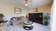 This single-floor residence offers a welcoming and well-appointed living space designed for comfort and functionality. The living room serves as the heart of the home, featuring plush wall-to-wall carpeting that adds warmth underfoot. Soft yellow walls create a bright, cheerful ambiance throughout the space, complemented by a textured ceiling and a classic ceiling fan with integrated lighting that ensures year-round comfort and practical illumination. A large dark wood entertainment center provides ample storage and display space, making it a natural focal point of the room. The seating area is anchored by a generously sized sofa in warm taupe tones, accented with decorative blue and grey throw pillows. A distinctive round coffee table with a glass top and upholstered seating panels adds both style and versatility to the arrangement. Decorative wall art and a framed mirror contribute to the room's curated, lived-in charm. The overall aesthetic blends casual comfort with thoughtful décor, resulting in a space that feels both inviting and functional — ideal for relaxing or entertaining.