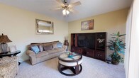 This single-floor residence offers a welcoming and well-appointed living space designed for comfort and functionality. The living room serves as the heart of the home, featuring plush wall-to-wall carpeting that adds warmth underfoot. Soft yellow walls create a bright, cheerful ambiance throughout the space, complemented by a textured ceiling and a classic ceiling fan with integrated lighting that ensures year-round comfort and practical illumination. A large dark wood entertainment center provides ample storage and display space, making it a natural focal point of the room. The seating area is anchored by a generously sized sofa in warm taupe tones, accented with decorative blue and grey throw pillows. A distinctive round coffee table with a glass top and upholstered seating panels adds both style and versatility to the arrangement. Decorative wall art and a framed mirror contribute to the room's curated, lived-in charm. The overall aesthetic blends casual comfort with thoughtful décor, resulting in a space that feels both inviting and functional — ideal for relaxing or entertaining.