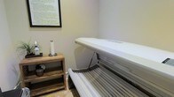This single-floor tanning space is designed with simplicity and functionality in mind, offering a clean and comfortable environment for personal tanning sessions. The room is finished with neutral-toned walls that create a calm, understated ambiance, complemented by light tile flooring that adds a tidy, polished feel underfoot.

The centerpiece of the space is a professional-grade tanning bed, positioned to make efficient use of the available area. Its sleek, modern design features a hinged canopy that opens to accommodate users comfortably, with integrated tanning lamps along both the top and bottom panels for an even, consistent session.

Adjacent to the tanning bed is a rustic, open-shelf wooden side table that provides convenient storage and surface space for personal items, towels, or accessories. A framed informational poster is mounted on the wall, likely providing safety guidelines and usage instructions, reflecting a commitment to client awareness and responsible tanning practices. A small decorative plant adds a subtle touch of nature to the room, softening the clinical feel of the equipment.

Overall, this tanning room balances practicality with a welcoming atmosphere, making it well-suited for use within a salon, wellness studio, or similar personal care facility.