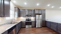 This single-floor home offers a clean, modern design with a cohesive aesthetic carried throughout every room. The kitchen is a standout feature, showcasing rich, dark grey cabinetry with raised panel detailing, light granite countertops, and a full suite of stainless steel appliances including a refrigerator, electric range, built-in microwave, and dishwasher. Recessed lighting illuminates the space, while a window allows natural light to filter in. A peninsula with a matching granite surface extends from the kitchen, creating a natural transition into the open living area and providing additional counter space.

The living and dining areas flow seamlessly from the kitchen, unified by warm hardwood-style flooring that runs throughout the home. Neutral, warm-toned walls paired with recessed lighting create a bright and inviting ambiance. Large windows bring in ample natural light, enhancing the open feel of the space.

The additional rooms maintain the same consistent flooring and neutral palette, offering versatile, light-filled spaces. The bathroom is finished with dark wood cabinetry, granite countertops, a wide vanity mirror, and a lighted fixture above, complementing the home's overall design language with both style and practicality.