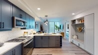 This single-floor residence showcases a thoughtfully designed interior with a contemporary aesthetic throughout. The kitchen serves as the focal point, featuring striking navy-blue shaker-style cabinetry extending nearly to the ceiling, paired with light quartz countertops and a matching marble-look tile backsplash. Stainless steel appliances, including a gas range, dishwasher, and built-in microwave, provide a professional-grade cooking environment. A central island with darker charcoal cabinetry offers additional workspace and open shelving, illuminated by a trio of modern pendant lights. Floor-to-ceiling windows at the far end of the kitchen and living area frame expansive urban skyline views, flooding the space with natural light.

Light wood-tone flooring runs consistently throughout, enhancing the open, airy feel. A built-in mudroom-style nook near the entry provides practical storage, while a clean, bright entryway with recessed lighting offers a welcoming arrival. The bathroom features bold hexagonal tile flooring, a dual-sink vanity with wood-toned cabinetry, a glass-enclosed walk-in shower, and bright white walls. The bedroom is comfortably scaled, featuring a ceiling fan and access to natural light.