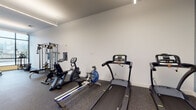 This modern fitness center offers a spacious, single-floor layout designed to support a full range of workout routines. The interior is finished with clean white walls, durable rubber flooring, and sleek linear pendant lighting that creates a bright, energizing atmosphere throughout the space. Exposed concrete columns add an industrial architectural character, while large floor-to-ceiling windows along one side of the room flood the space with natural light and provide views to the outdoors.

The cardio area is well-stocked with multiple treadmills, rowing machines, stationary bikes — including air bikes — and elliptical trainers, offering ample options for cardiovascular training. A dedicated strength training section features a multi-station cable and weight machine, a flat bench press, and a rack of dumbbells and kettlebells in varying sizes, catering to both beginners and experienced lifters. A functional fitness zone includes a rack stocked with medicine balls, resistance bands, and exercise mats, providing versatile options for dynamic and flexibility-focused training. Wall-mounted mirrors in key areas enhance the sense of space and allow users to monitor their form. The overall layout is thoughtfully organized to allow comfortable movement between equipment zones, making this fitness center a well-rounded and accessible facility.