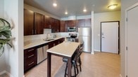 This thoughtfully designed one-bedroom, one-bathroom residence presents a cohesive and functional single-floor layout that balances modern style with everyday comfort. The kitchen serves as a standout feature, showcasing rich dark-wood cabinetry, light quartz countertops, a classic subway tile backsplash, and a full suite of stainless steel appliances, including a range, microwave, dishwasher, and refrigerator. A central island with bar seating provides additional prep space and a casual dining option, making the kitchen both practical and social. Light wood-style flooring flows through the kitchen and living areas, creating a warm and cohesive aesthetic. The living room offers a welcoming atmosphere with neutral tones, recessed lighting, and large windows fitted with vertical blinds that allow for natural light control. The bedroom is a serene retreat featuring plush carpeting, a broad window that fills the room with natural light, and access to a walk-in closet for generous storage. The bathroom is cleanly appointed with dark wood vanity cabinetry, a white countertop, a framed mirror, and a shower-tub combination enclosed by a light striped curtain. Tile-style flooring completes the bathroom's polished look. Overall, the residence presents a modern, move-in-ready environment with a neutral palette and quality finishes throughout.