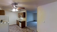 This single-floor residence offers a practical and straightforward layout with a seamless flow between the kitchen and living areas. The kitchen is equipped with warm-toned wood cabinetry providing ample upper and lower storage, a countertop with an integrated sink, and a dishwasher. A ceiling fan with a built-in light fixture keeps the kitchen area well-lit and ventilated. The flooring in the kitchen features a durable tile pattern, adding a functional contrast to the space. Adjacent to the kitchen, the open living area transitions to plush carpeting, creating a comfortable and distinct zone for relaxation. A baseboard heating unit runs along the far wall of the living area, ensuring efficient climate control throughout the cooler months. The walls throughout are finished in neutral tones, offering a clean and versatile backdrop. The textured ceiling adds a subtle architectural detail consistent across both spaces. The open sightline between the kitchen and living room enhances the overall sense of space, making the layout feel connected and easy to navigate.