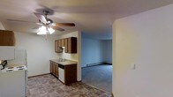 This single-floor residence offers a practical and straightforward layout with a seamless flow between the kitchen and living areas. The kitchen is equipped with warm-toned wood cabinetry providing ample upper and lower storage, a countertop with an integrated sink, and a dishwasher. A ceiling fan with a built-in light fixture keeps the kitchen area well-lit and ventilated. The flooring in the kitchen features a durable tile pattern, adding a functional contrast to the space. Adjacent to the kitchen, the open living area transitions to plush carpeting, creating a comfortable and distinct zone for relaxation. A baseboard heating unit runs along the far wall of the living area, ensuring efficient climate control throughout the cooler months. The walls throughout are finished in neutral tones, offering a clean and versatile backdrop. The textured ceiling adds a subtle architectural detail consistent across both spaces. The open sightline between the kitchen and living room enhances the overall sense of space, making the layout feel connected and easy to navigate.