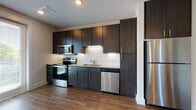 This contemporary single-floor residence blends modern design with functional living spaces. The kitchen is a standout feature, outfitted with dark espresso cabinetry that extends to the ceiling for maximum storage, granite countertops, a white subway tile backsplash, and a full suite of stainless steel appliances including a range, microwave, dishwasher, and refrigerator. Under-cabinet lighting adds a polished touch, while warm hardwood-style flooring ties the space together seamlessly.

The living area is bright and welcoming, featuring large French doors flanked by a full-length sidelight window that invites an abundance of natural light and provides direct outdoor access. The open layout flows naturally, enhanced by neutral wall tones and clean architectural lines.

The bedrooms are carpeted for comfort, each offering generous natural light through large windows with views of surrounding greenery. One bedroom includes a ceiling fan for added comfort. Built-in shelving in the living area provides convenient display and storage options.

The bathroom is neatly appointed with a soaking tub and shower combination with white tile surround, hardwood-style flooring, a granite-topped vanity, and dark cabinetry consistent with the kitchen's design language. Overall, the space presents a cohesive, modern aesthetic throughout.