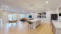 Located at 18217 Cary Street in Omaha, Nebraska, this thoughtfully designed single-floor space seamlessly integrates a kitchen, dining room, and living room into one cohesive, open-plan layout. Light hardwood flooring flows throughout, complementing the crisp white walls and clean-lined modern aesthetic.

The kitchen serves as a central hub, anchored by a substantial white quartz island with seating for multiple guests. Stainless steel appliances — including a full-size refrigerator, microwave, and dishwasher — line the cabinetry, while elegant glass pendant lights hang overhead. A wall-mounted flat-screen television adds a functional, entertainment-ready element to the space.

Adjacent to the kitchen, the dining area features a warm wood communal table surrounded by contemporary chairs. Floor-to-ceiling glass doors open to an outdoor deck, flooding the room with natural light and providing views of the surrounding area. Framed artwork adds curated character to the walls.

The living room offers a comfortable retreat with two tufted cognac leather sofas, a round coffee table, and additional seating. Windows with horizontal blinds frame outdoor views, while a wall-mounted television and recessed lighting enhance the room's inviting ambiance. The overall space strikes a balance between functionality and modern style, making it well-suited for both casual gatherings and everyday use.