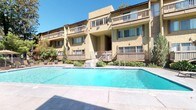 Hidden Springs is a well-appointed residential community designed with both comfort and functionality in mind. The exterior showcases a warm stucco facade with multiple levels of private balconies overlooking lush landscaping and a large, sparkling outdoor pool. The pool area is framed by a spacious tile deck, manicured greenery, and easy access to the interior clubhouse, creating a seamless indoor-outdoor living experience.

The clubhouse interior features soaring ceilings with transom windows that flood the space with natural light and frame direct views of the pool. Large French doors open onto the pool deck, blurring the boundary between inside and out. The entry area is finished with handsome slate tile flooring, which continues throughout the main living space, lending the interior a grounded, earthy aesthetic.

The lounge area offers comfortable seating arranged around a central coffee table, ideal for relaxed gatherings. A dedicated office nook, branded with the Hidden Springs name, provides a workspace with a full desk setup and leather seating. A graceful staircase with decorative wrought-iron railings connects the main floor to upper levels, adding an elegant architectural focal point. Recessed lighting and warm wall tones throughout contribute to the inviting, community-centered ambiance of the space.