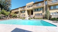 Hidden Springs is a well-appointed residential community designed with both comfort and functionality in mind. The exterior showcases a warm stucco facade with multiple levels of private balconies overlooking lush landscaping and a large, sparkling outdoor pool. The pool area is framed by a spacious tile deck, manicured greenery, and easy access to the interior clubhouse, creating a seamless indoor-outdoor living experience.

The clubhouse interior features soaring ceilings with transom windows that flood the space with natural light and frame direct views of the pool. Large French doors open onto the pool deck, blurring the boundary between inside and out. The entry area is finished with handsome slate tile flooring, which continues throughout the main living space, lending the interior a grounded, earthy aesthetic.

The lounge area offers comfortable seating arranged around a central coffee table, ideal for relaxed gatherings. A dedicated office nook, branded with the Hidden Springs name, provides a workspace with a full desk setup and leather seating. A graceful staircase with decorative wrought-iron railings connects the main floor to upper levels, adding an elegant architectural focal point. Recessed lighting and warm wall tones throughout contribute to the inviting, community-centered ambiance of the space.