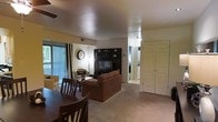This single-floor residence offers a thoughtfully arranged open-concept layout that seamlessly connects the dining and living areas, creating a cohesive and functional living space. The living room is anchored by a dark, built-in entertainment wall unit with shelving, providing ample storage and display space alongside a central television. A plush brown sofa and coordinating armchair complete the seating arrangement, while soft carpeted flooring runs throughout, adding warmth and comfort underfoot. A ceiling fan overhead enhances airflow throughout the shared living space. The dining area is positioned adjacent to the living room and features a dark wood dining table with matching chairs, ideal for everyday meals or casual entertaining. Natural light flows in generously through a large sliding glass door dressed with vertical blinds, which opens to an outdoor area and provides a pleasant connection to the exterior. A hallway visible from the main living area leads to additional rooms, including what appears to be a laundry area, while a set of double doors near the entryway suggests a spacious closet for extra storage. The overall ambiance is comfortable, practical, and well-suited for everyday living.