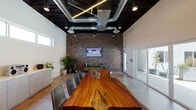 This single-floor commercial space blends industrial and contemporary design elements to create a distinctive and functional meeting environment. The centerpiece of the room is a large live-edge wood conference table, showcasing natural grain and organic contours, surrounded by sleek gray leather chairs. The dramatically dark ceiling features exposed ductwork, linear pendant lighting, and recessed spotlights, creating a bold industrial contrast against the crisp white walls.

A full-height brick accent wall anchors one end of the room, hosting a wall-mounted display screen and flanked by greenery, adding warmth and texture to the space. Opposite, expansive glass sliding doors flood the interior with natural light and provide direct access to an outdoor courtyard lined with planters and seating. A continuous band of clerestory windows along the upper walls further enhances natural light throughout.

A built-in credenza with light wood cabinetry lines one wall, offering functional storage and display space. A separate utility area includes a double-basin sink with brass fixtures, light wood cabinetry, and white tile backsplash. Polished concrete floors tie the space together, reinforcing the clean, modern-industrial aesthetic throughout.