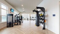 Located at 3785 Bismarck Ct in Wildwood, Florida, this single-story fitness center is designed to deliver a comprehensive workout experience in a bright, contemporary setting. The space is finished with striking herringbone-patterned light wood flooring throughout, lending a clean and modern aesthetic that complements the crisp white walls and recessed lighting overhead. Ceiling fans are strategically placed to ensure comfortable air circulation during workouts.

Large, multi-pane windows line multiple walls, flooding the interior with abundant natural light and offering views of the surrounding outdoor pool area and landscaped grounds. The open layout is spacious enough to accommodate a wide variety of fitness zones without feeling crowded. Cardio equipment includes treadmills, ellipticals, stationary and spin bikes, and a rowing machine. Strength training is well represented with cable machines, a multi-station functional trainer, a dedicated free weights and dumbbell rack, and a heavy bag suspended from a freestanding rig.

A mirrored wall anchors one section of the facility, ideal for stretching or bodyweight training, while wall-mounted televisions are positioned throughout for entertainment. Plyo boxes and additional accessories round out the functional training offerings, making this fitness center a versatile and well-appointed space for a full range of exercise routines.