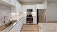 This well-appointed single-floor residence offers a thoughtfully designed open-concept layout that seamlessly connects the kitchen and living areas. The kitchen is a standout feature, showcasing crisp white shaker-style cabinetry, granite countertops with subtle veining, a classic subway tile backsplash, and a full suite of stainless steel appliances including a refrigerator, range, and built-in microwave. A central kitchen island with a matching granite surface and pendant lighting above serves as both a functional workspace and a natural gathering point, opening directly into the spacious living area. Warm, light-toned wood-look flooring flows throughout the main living spaces, contributing to a clean and contemporary aesthetic. The bedroom is carpeted for comfort, features neutral walls, a ceiling fan with integrated lighting, and a window that allows natural light to filter in through blinds. The bathroom includes a full tub and shower combo with a curved curtain rod, clean white tile surround, and wood-look flooring, accompanied by an adjacent walk-in closet with multi-tiered shelving and hanging storage. The overall design palette of whites, soft grays, and warm wood tones creates a cohesive, modern ambiance throughout the space.