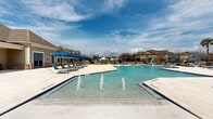 This resort-style outdoor amenity space centers around a generously sized swimming pool with crystal-clear blue water and a shallow entry area, making it accessible and inviting. Multiple fountain jets rise from the pool's surface, adding a dynamic visual element to the overall ambiance. The pool is flanked by an expansive travertine-tiled sun deck furnished with rows of cushioned lounge chairs and coordinating blue market umbrellas, providing ample space for sunbathing and outdoor leisure.

A standout architectural feature of the space is the open-air pavilion, constructed with stone columns, exposed wood timber framing, and a metal standing-seam roof. This covered structure houses additional seating and gathering areas, offering a shaded retreat while maintaining an open, airy feel. A stone fireplace anchors one end of the pavilion, enhancing its appeal during cooler seasons.

Adjacent to the pavilion sits a clubhouse-style building with large windows and traditional architectural detailing, complementing the overall design of the community. The pool area is enclosed by a black wrought-iron fence, ensuring privacy and security. Palm trees and landscaped plantings are thoughtfully positioned throughout, contributing to the tropical, resort-like atmosphere of this well-appointed outdoor space.