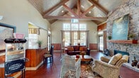 Located at 5799 Southland Dr in Mobile, Alabama, this single-floor community space showcases a compelling blend of rustic charm and welcoming design. The centerpiece of the main hall is a dramatic vaulted ceiling adorned with exposed wood trusses and tongue-and-groove wood planking, soaring overhead to create an impressive sense of openness and volume. Rich hardwood floors run throughout, complementing the warm wood wainscoting that lines the lower walls.

A floor-to-ceiling stacked stone fireplace anchors one end of the main gathering area, serving as a striking architectural focal point. Multiple seating arrangements — including leather sofas, wicker chairs, and upholstered armchairs — create inviting conversation areas beneath the cathedral ceiling. Large multi-pane windows flood the interior with abundant natural light, offering views of the surrounding landscape.

A built-in bar counter with granite surfaces and bar-height seating provides a functional area for refreshments, while an adjacent kitchenette nook offers a coffee station, mini-refrigerator, and casual dining seating with eclectic wall art. French doors with glass panels lead to additional office or multipurpose rooms, and exterior glass doors open to an outdoor area. The overall ambiance is warm, inviting, and distinctly lodge-inspired.