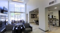 This single-floor space makes a striking impression through its dramatic double-height ceilings and expansive floor-to-ceiling windows that flood the interior with natural light and provide open views to the surrounding outdoor areas. The main lounge area is anchored by plush, tufted gray sofas accented with blue and white decorative pillows, complemented by sleek leather armchairs and a dark accent coffee table. A statement geometric chandelier hangs overhead, adding a refined focal point to the soaring ceiling.

The walls are adorned with an curated gallery of decorative mirrors and framed artwork mounted against dark accent panels, adding visual depth and sophistication to the bright white surroundings. Large-format tile flooring in a neutral tone runs throughout, contributing to a clean, cohesive aesthetic.

An adjacent dining area features a round table with contemporary seating, while a well-appointed kitchen with dark cabinetry, pendant lighting, and stainless steel appliances is visible through an open archway. Glass entry doors at the far end enhance the sense of openness, connecting the interior seamlessly to the outside.