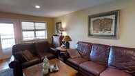 This single-floor space centers around a comfortable and well-appointed living room that exudes a warm, welcoming ambiance. The room is furnished with rich, dark brown leather seating — including a sofa and matching armchair — arranged around a solid wood coffee table, creating an intimate and functional seating area. Warm neutral tones on the walls complement the earthy color palette throughout. A wooden end table holds a classic table lamp, casting a soft, warm glow that enhances the room's cozy atmosphere. Natural light filters in through a window dressed with a neutral roller shade, adding brightness to the space. A wooden dresser or chest of drawers serves as both storage and a display surface, adding a traditional character to the room. The walls are thoughtfully decorated with framed artwork, including a prominently displayed map-style print in a gold frame, lending a cultured and personal touch to the interior. Recessed ceiling lighting provides additional illumination, ensuring the space is well-lit at all times. Overall, this space offers a comfortable, lived-in aesthetic with a balance of classic furnishings and personal touches that make it feel distinctly homey and inviting.