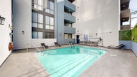 Located at 1223 Federal Ave in the heart of West Los Angeles, this outdoor pool courtyard offers a well-appointed amenity space designed for relaxation and recreation. The centerpiece is a generously sized rectangular swimming pool with crystal-clear turquoise water, accessible via stainless steel handrails on multiple sides. The pool deck is finished in smooth concrete and lined with sleek lounge chairs, providing ample space for sunbathing and unwinding. A dedicated seating area with wicker furniture offers a comfortable spot to gather or relax poolside. The courtyard is enclosed on multiple sides by the surrounding multi-story building, which features a contemporary facade of white stucco and blue accent panels, large grid-pane windows, and tiered balconies that overlook the pool area. A vertical green wall adds a natural, vibrant contrast to the architectural elements. The open-air design allows natural light to filter into the courtyard throughout the day. Additional amenities visible in the space include wall-mounted pool equipment and signage. The overall ambiance is clean, modern, and functional, reflecting a well-maintained residential community in a prime Los Angeles location.