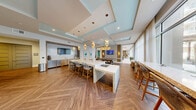Located in Winter Park, Florida, this thoughtfully designed amenity space combines entertainment and productivity in a single, open-plan layout. The game room anchors the space with a full-size billiard table positioned beneath a striking sculptural gold pendant light, complemented by herringbone-patterned hardwood flooring that flows throughout. Coffered ceilings with soft teal accents add architectural depth and a sense of height, while floor-to-ceiling windows invite abundant natural light and offer views of lush palm-lined outdoor surroundings. A built-in wet bar area features warm wood cabinetry, a wall-mounted flat-screen TV, a microwave, and a sink — ideal for casual gatherings. A long quartz-topped communal island with bar-height seating serves as a central hub for collaboration or socializing. Along one wall, a row of curved teal banquette booths with organic, wave-shaped dividers creates intimate lounge nooks, each lit by sculptural pendant lights and paired with tulip-style tables and upholstered stools. The adjacent office area provides semi-private workstations with counter seating, maintaining the same cohesive coastal-modern aesthetic. Curated teal abstract artwork and carefully layered lighting complete the polished, resort-inspired atmosphere of this dynamic space.
