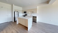 This single-floor unit offers a clean, contemporary design with a thoughtful layout that maximizes both functionality and comfort. The open-concept kitchen is a standout feature, showcasing light wood cabinetry, a large central island with a white quartz countertop, and a full suite of stainless steel appliances including a refrigerator, range, microwave, and dishwasher. Light wood-tone flooring flows throughout the kitchen and living areas, contributing to a bright, airy atmosphere. The living area is generously proportioned, featuring large windows and a glass door that open to an outdoor balcony, flooding the space with natural light and offering scenic views of surrounding greenery. A wall-mounted climate control unit ensures year-round comfort. The bedroom continues the light and neutral palette, featuring plush carpeting, a ceiling fan, and a large window with expansive outdoor views, creating a peaceful and restful environment. The bathroom is efficiently designed with a wide light wood vanity, white quartz countertop, an undermount sink, and a shower-tub combination. The unit also includes an in-unit laundry area, as visible in the floor plan. Overall, the space presents a modern, move-in-ready environment with a cohesive neutral aesthetic throughout.