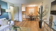 This single-floor residence showcases a thoughtfully designed open-concept layout that connects the living, dining, and kitchen areas in a natural, flowing arrangement. Rich hardwood flooring extends throughout the main living spaces, adding warmth and visual continuity to the interior.

The dining area serves as a focal point of the space, highlighted by a bold accent wall in a warm amber tone that draws the eye and creates a welcoming atmosphere. A round dark-wood dining table surrounded by upholstered chairs sits comfortably beneath a track lighting fixture, providing both functional illumination and a stylish finishing touch.

The kitchen is positioned at the rear of the open living area and features white cabinetry, dark countertops, and ample storage. The white cabinetry contrasts cleanly with the warmer tones of the surrounding space, lending a crisp, organized feel.

A textured ceiling adds subtle architectural character throughout the main area, while neutral wall tones provide a versatile backdrop that complements both the furnishings and natural accents. Decorative greenery and floral arrangements are used to soften the space and introduce natural elements. Multiple doorways visible throughout suggest a well-connected and practical floor plan. Overall, the space exudes a cozy yet polished ambiance suited to everyday living and entertaining alike.