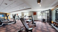This fitness center is thoughtfully designed to support a wide range of workout routines across two distinct zones. The main cardio and strength training area is anchored by patterned carpet flooring, bold accent walls in red and orange, and a generous array of equipment including treadmills, elliptical trainers, stationary bikes, cable machines, and free weights. A built-in media cabinet with a wall-mounted television serves as a focal point in this area, while recessed and overhead lighting ensures excellent visibility throughout. Multiple large windows line the perimeter, allowing natural light to fill the space and offering views of the surrounding greenery.

Adjoining the main floor is a dedicated studio room featuring light hardwood-style flooring, a large wall-mounted display screen ideal for guided fitness classes, and a chalkboard accent wall that adds a dynamic, interactive element. This versatile space accommodates rowing machines, spin bikes, and open floor stretching, with ceiling fans providing additional comfort. Decorative sculptural wall art and a full-length mirror enhance the studio's modern aesthetic. Together, both areas create a comprehensive, energizing fitness environment suited for cardiovascular training, strength conditioning, and group exercise.