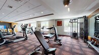This fitness center is thoughtfully designed to support a wide range of workout routines across two distinct zones. The main cardio and strength training area is anchored by patterned carpet flooring, bold accent walls in red and orange, and a generous array of equipment including treadmills, elliptical trainers, stationary bikes, cable machines, and free weights. A built-in media cabinet with a wall-mounted television serves as a focal point in this area, while recessed and overhead lighting ensures excellent visibility throughout. Multiple large windows line the perimeter, allowing natural light to fill the space and offering views of the surrounding greenery.

Adjoining the main floor is a dedicated studio room featuring light hardwood-style flooring, a large wall-mounted display screen ideal for guided fitness classes, and a chalkboard accent wall that adds a dynamic, interactive element. This versatile space accommodates rowing machines, spin bikes, and open floor stretching, with ceiling fans providing additional comfort. Decorative sculptural wall art and a full-length mirror enhance the studio's modern aesthetic. Together, both areas create a comprehensive, energizing fitness environment suited for cardiovascular training, strength conditioning, and group exercise.