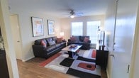 This single-floor space offers a comfortable and thoughtfully designed living environment. The living room serves as the centerpiece, featuring rich hardwood flooring that adds warmth and character throughout. Two dark leather sofas are arranged around a sleek glass-topped coffee table, all anchored by a bold geometric area rug in shades of red, black, gray, and white that brings a contemporary flair to the room. A ceiling fan with an integrated light fixture is centrally positioned, providing both air circulation and ambient lighting. Natural light pours in through multiple windows fitted with white blinds, creating a bright and airy atmosphere. The walls are decorated with framed artwork, adding a personalized touch to the neutral palette. A large flat-screen television is mounted on a media console, making the space well-suited for both relaxation and entertainment. Accent pillows in red and patterned designs complement the color scheme, tying the room's aesthetic together cohesively. The overall ambiance is modern, clean, and inviting, with a balanced mix of comfort and style that makes the living area both functional and visually appealing.
